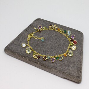 May include: A gold chain bracelet with a variety of colorful gemstones dangling from it. The bracelet is on a gray stone surface.