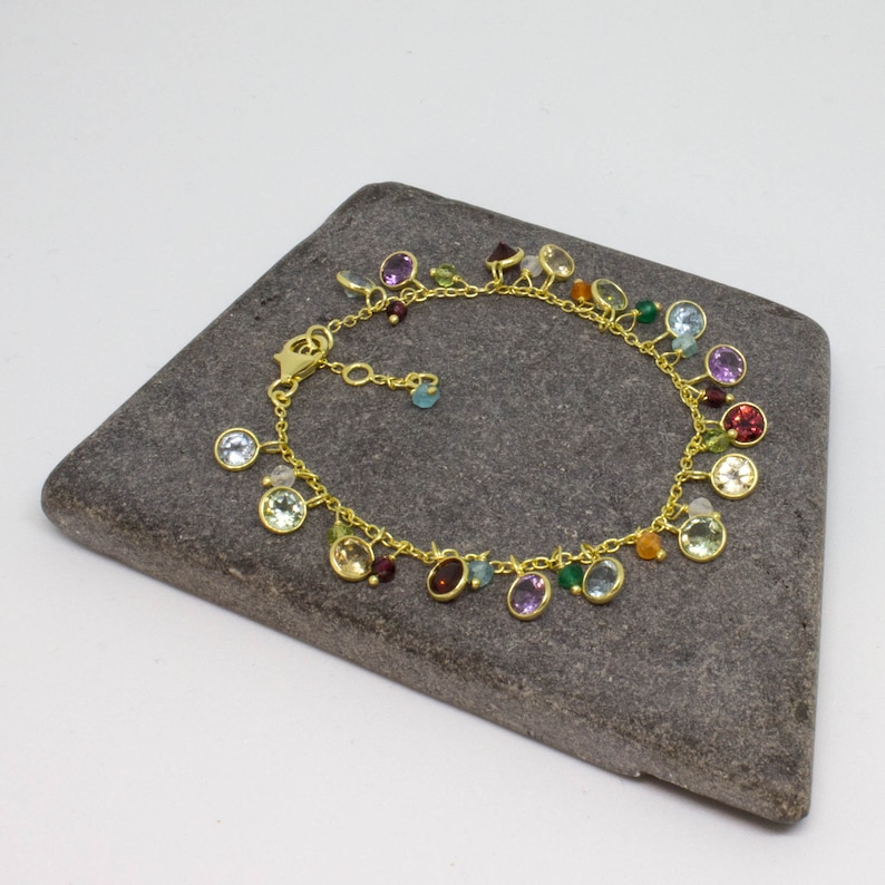 May include: A gold chain bracelet with a variety of colorful gemstones dangling from it. The bracelet is on a gray stone surface.