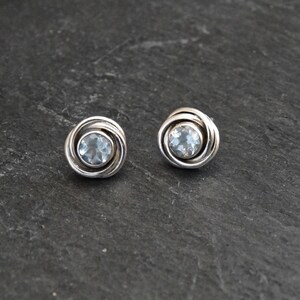 May include: A pair of silver earrings with a light blue gemstone in the center of each earring. The earrings are round and have a twisted design.