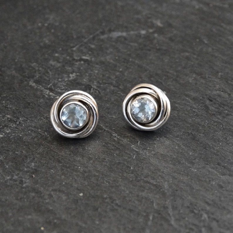 May include: A pair of silver earrings with a light blue gemstone in the center of each earring. The earrings are round and have a twisted design.