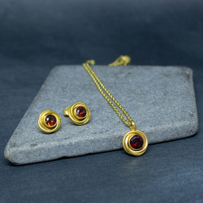 Op de afbeelding: Een gouden ketting en oorbellen set met een rode edelsteen hanger en bijpassende oorbellen. De sieraden worden getoond op een grijs stenen oppervlak.