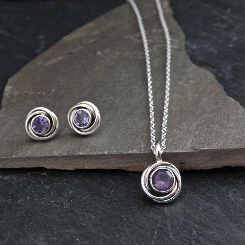 K&ouml;nnte beinhalten: Ein silbernes Halsketten- und Ohrring-Set mit einem violetten Amethyststein in einem runden Anh&auml;nger- und Ohrstecker-Design.