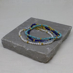 May include: Four beaded bracelets stacked on a gray stone surface. The bracelets are made of white pearls, light blue, turquoise, and dark blue beads. Each bracelet has a gold clasp.