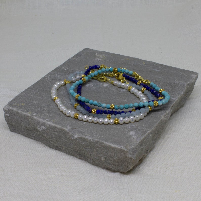 May include: Four beaded bracelets stacked on a gray stone surface. The bracelets are made of white pearls, light blue, turquoise, and dark blue beads. Each bracelet has a gold clasp.