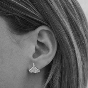 May include: A pair of silver earrings shaped like ginkgo leaves. The earrings are small and delicate, and they are worn in the wearer's earlobes.