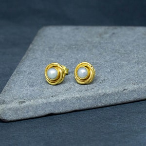 May include: A pair of gold earrings with a white pearl in the center of each earring. The earrings are on a gray surface.
