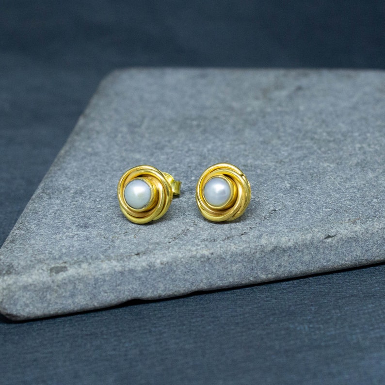 May include: A pair of gold earrings with a white pearl in the center of each earring. The earrings are on a gray surface.