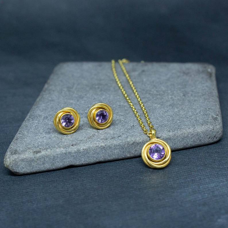K&ouml;nnte beinhalten: Ein Gold-Halsketten- und Ohrring-Set mit einem violetten Edelstein in jedem St&uuml;ck. Die Halskette hat eine zarte Kette und einen Anh&auml;nger mit einem runden Design. Die Ohrringe sind klein und haben ein &auml;hnliches rundes Design.