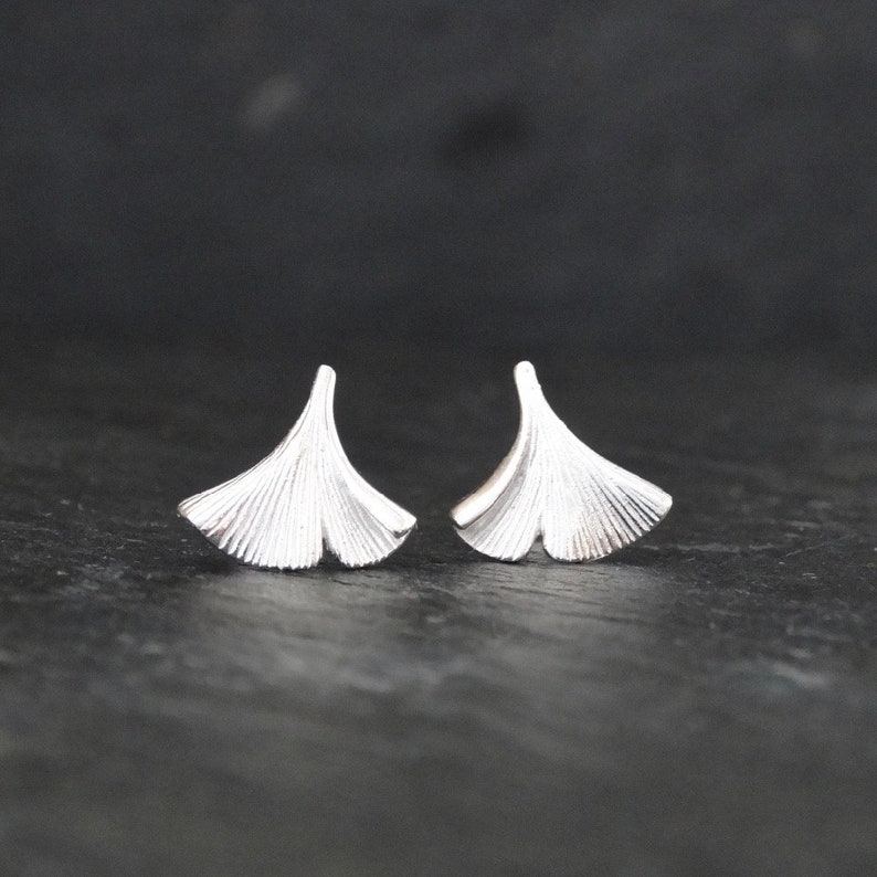 May include: A pair of silver ginkgo leaf stud earrings. The earrings are small and delicate, and they have a textured surface.