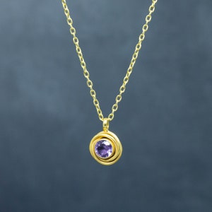 K&ouml;nnte beinhalten: Eine goldene Kettenkette mit einem Anh&auml;nger, der einen violetten Edelstein zeigt, der von einem goldenen Wirbelmuster umgeben ist.