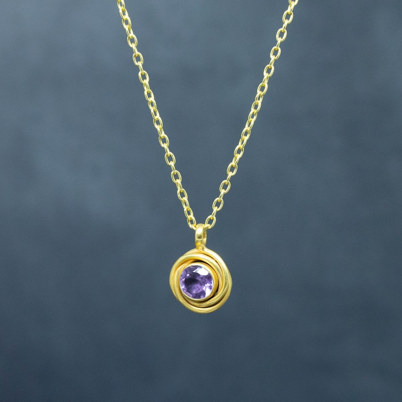 K&ouml;nnte beinhalten: Eine goldene Kettenkette mit einem Anh&auml;nger, der einen violetten Edelstein zeigt, der von einem goldenen Wirbelmuster umgeben ist.