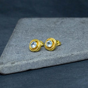 May include: A pair of gold earrings with a light blue gemstone in the center of each earring. The earrings are on a gray surface.