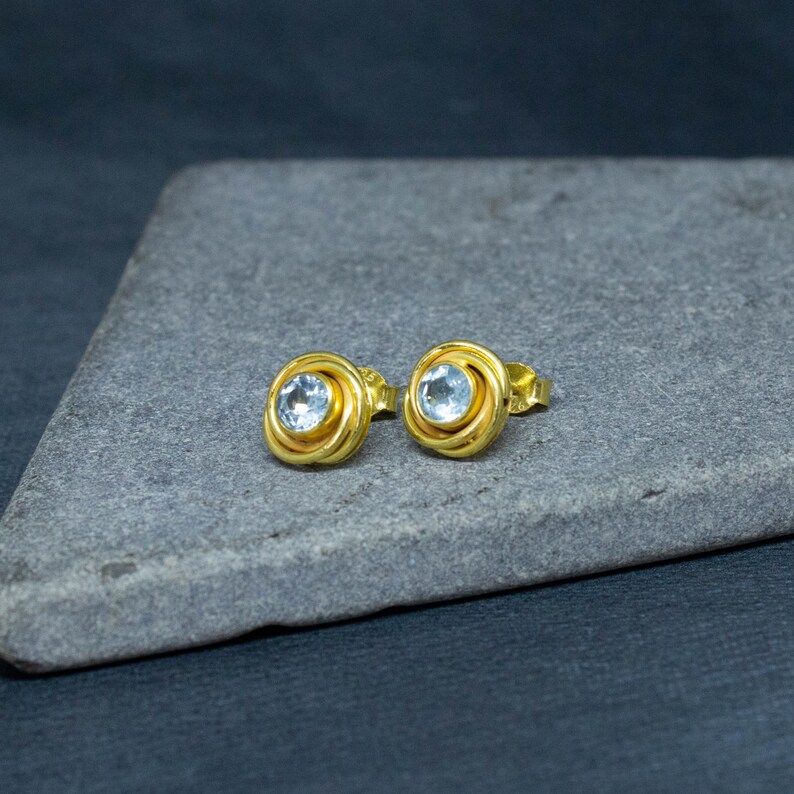May include: A pair of gold earrings with a light blue gemstone in the center of each earring. The earrings are on a gray surface.