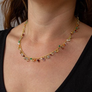 May include: A gold chain necklace with a variety of colorful gemstones dangling from it. The necklace is worn by a person with a black shirt.