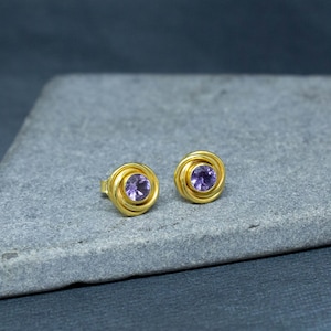 K&ouml;nnte beinhalten: Ein Paar goldene Ohrstecker mit einem violetten Amethyst-Edelstein in der Mitte jedes Ohrsteckers.