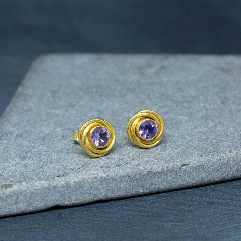 K&ouml;nnte beinhalten: Ein Paar goldene Ohrstecker mit einem violetten Amethyst-Edelstein in der Mitte jedes Ohrsteckers.