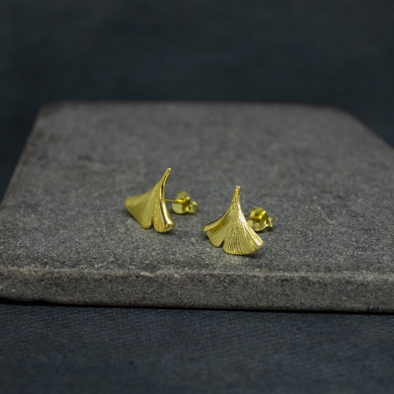 May include: A pair of gold-toned earrings shaped like ginkgo leaves. The earrings are on a gray stone surface.