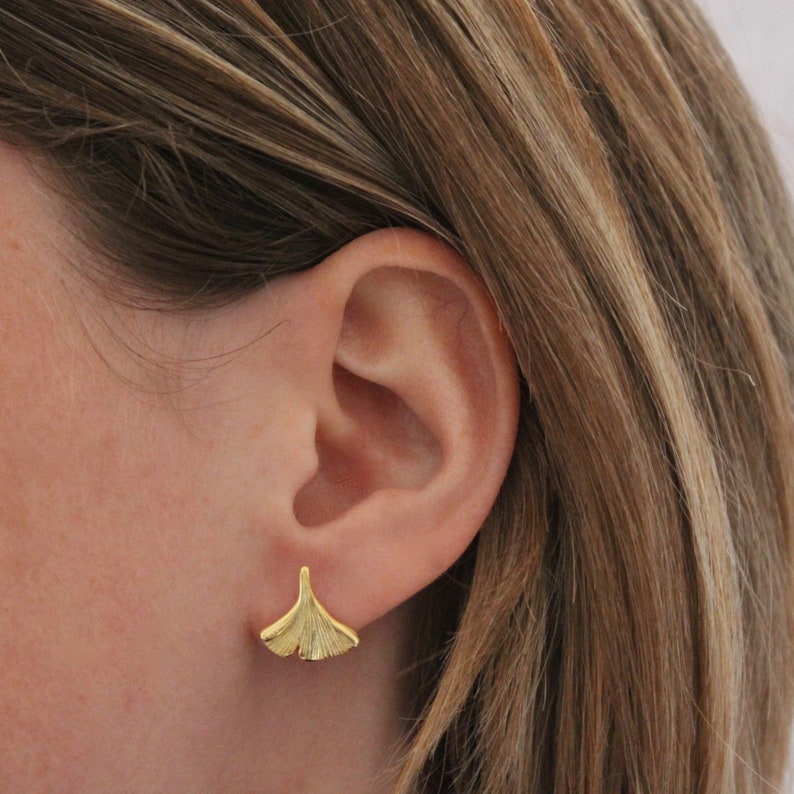 May include: A pair of gold-toned ginkgo leaf earrings. The earrings are small and delicate, and they have a simple, elegant design.