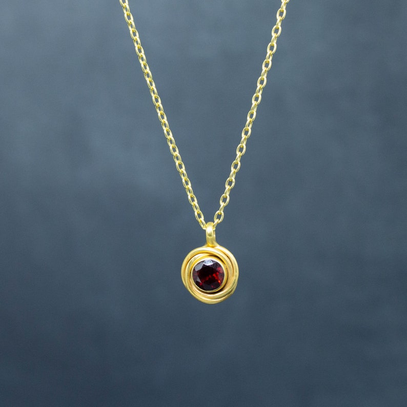 Op de afbeelding: Een gouden ketting met een hanger met een rode edelsteen omgeven door een gouden cirkel.