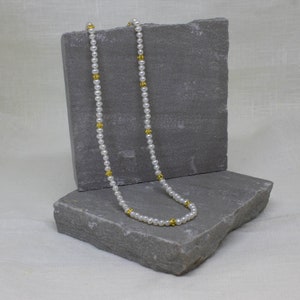 May include: A white pearl necklace with gold accents. The necklace is displayed on a gray stone surface.