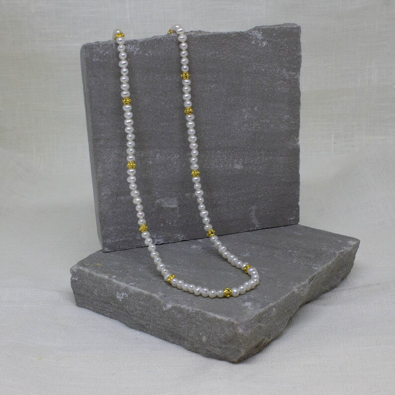 May include: A white pearl necklace with gold accents. The necklace is displayed on a gray stone surface.