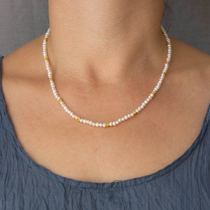 May include: A delicate white pearl necklace with small gold beads interspersed throughout. The necklace is worn by a person, but the focus is on the necklace itself.