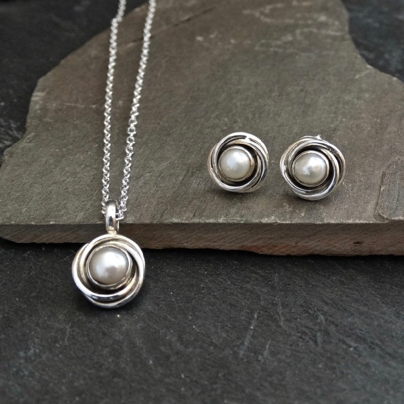 May include: A silver necklace and earrings set with a pearl in each piece. The pearl is set in a silver swirl design.