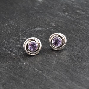 K&ouml;nnte beinhalten: Ein Paar silberne Ohrringe mit einem violetten Amethyst-Edelstein in der Mitte jedes Ohrrings. Die Ohrringe haben ein wirbelndes Design um den Edelstein herum.