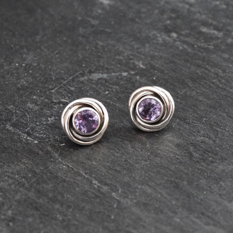 K&ouml;nnte beinhalten: Ein Paar silberne Ohrringe mit einem violetten Amethyst-Edelstein in der Mitte jedes Ohrrings. Die Ohrringe haben ein wirbelndes Design um den Edelstein herum.