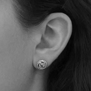 May include: A close-up of a person's ear with a silver earring featuring a round, clear gemstone surrounded by a silver swirl design.