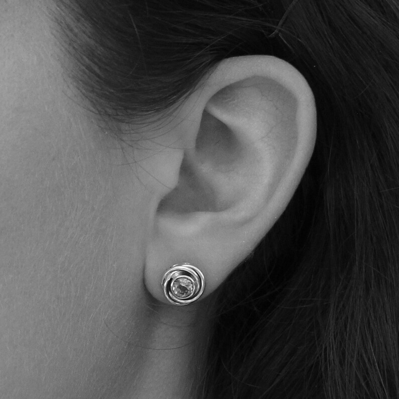 May include: A close-up of a person's ear with a silver earring featuring a round, clear gemstone surrounded by a silver swirl design.