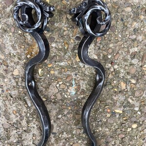 Op de afbeelding: Twee zwarte metalen deurgrepen met een drakenkopontwerp. De grepen zijn gebogen en hebben een decoratieve ring aan de bovenkant.