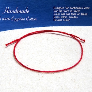 May include: A thin red string bracelet made of 100% Egyptian cotton. The text reads: Handmade with 100% Egyptian Cotton. Designed for continuous wear. Can be worn in water. Color will not fade or bleed. Dries within minutes. Retains luster.