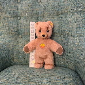May include: A vintage-style teddy bear in a light brown color, with darker brown accents on the paws and ears. The bear has a small, round, yellow and red tag on its chest. A ruler is placed next to the bear, measuring up to 12 inches.