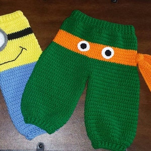 May include: Two pairs of crocheted baby pants. One pair is yellow and blue with a Minion face on the front. The other pair is green with an orange stripe and two white eyes.
