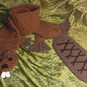 May include: A brown crocheted beaver scarf with a tail, head, and paws. The scarf is designed to be worn around the neck with the head and tail hanging down.