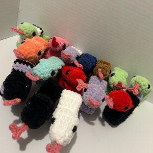 May include: A collection of colorful, crocheted axolotl plushies. The axolotls are in various colors, including black, white, red, orange, green, and purple. Each plushie has small black eyes and pink gills and feet.