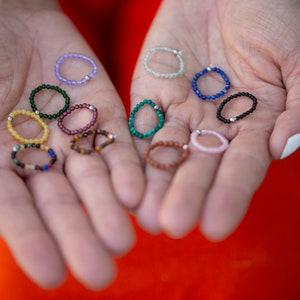 Puede incluir: Una colección de anillos de cuentas de colores, cada uno con un acento de cuentas de plata. Los anillos están dispuestos en las palmas abiertas de una persona.