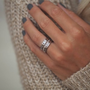 May include: A close-up of a hand wearing three silver rings. The rings are stacked on each other and have engraved words on them. The top ring says "Shea", the middle ring has a heart symbol, and the bottom ring says "Kadi".