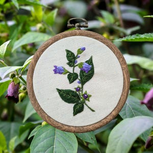 May include: Embroidered hoop art featuring a purple flower with green leaves and purple berries. The hoop is made of wood and has a brown finish.