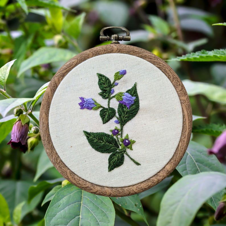 May include: Embroidered hoop art featuring a purple flower with green leaves and purple berries. The hoop is made of wood and has a brown finish.