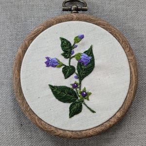 May include: Embroidered hoop art featuring a purple flower with green leaves and purple berries. The hoop is made of wood and has a natural finish.