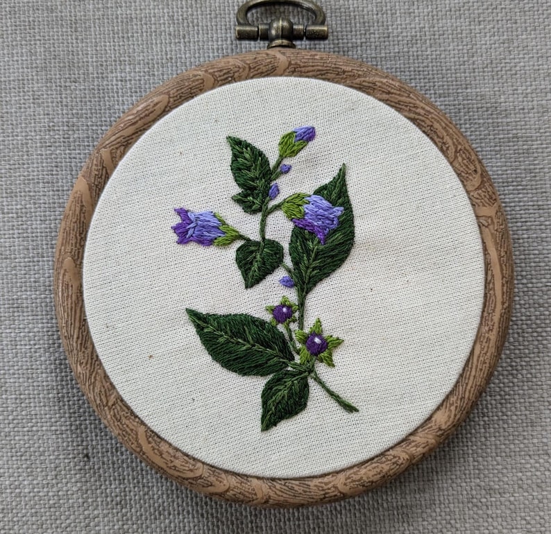 May include: Embroidered hoop art featuring a purple flower with green leaves and purple berries. The hoop is made of wood and has a natural finish.
