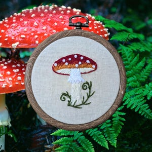 May include: Embroidered mushroom art in a wooden hoop. The mushroom has a red cap with white spots, an orange underside, and a white stem. Green fern and a red mushroom are in the background.