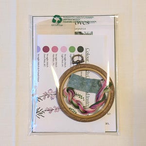 May include: A close-up of an embroidery kit with a wooden hoop, floss, and instructions. The kit includes four colours of floss: light green, light pink, mid pink, and dark pink. The instructions include a colour chart and a list of stitches.