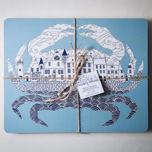 May include: Two blue and white crab-shaped coasters with a town scene inside the crab's body. The coasters are tied together with twine and a tag that reads "Crocky Neuk, Whistercross, www.crockyneuk.com".