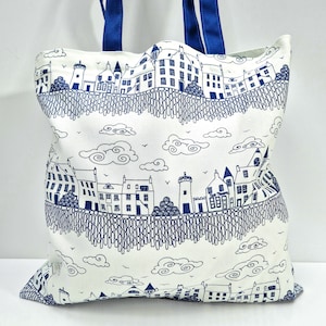 Puede incluir: Un bolso tote blanco con una ilustraci&oacute;n azul y blanca de un paisaje urbano. El dise&ntilde;o presenta edificios, nubes y una estructura similar a una valla. El bolso tiene asas azules.