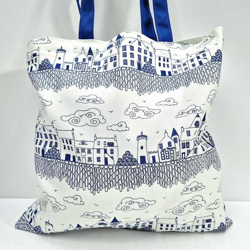 Puede incluir: Un bolso tote blanco con una ilustraci&oacute;n azul y blanca de un paisaje urbano. El dise&ntilde;o presenta edificios, nubes y una estructura similar a una valla. El bolso tiene asas azules.