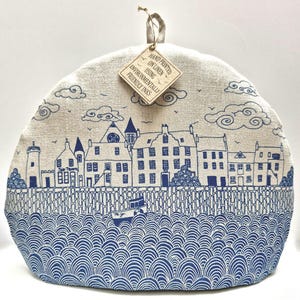 May include: A beige tea cozy with a blue coastal town design. The design includes a lighthouse, buildings, a boat, and stylized waves. The tag reads "HAND PRINTED ON LINEN USING ENVIRONMENTALLY FRIENDLY INKS."