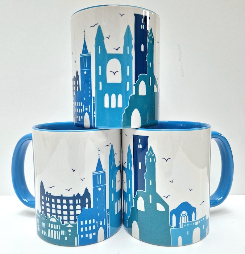 May include: Three blue and white ceramic mugs with a cityscape design. Each mug features a different silhouette of a city skyline with buildings and birds.